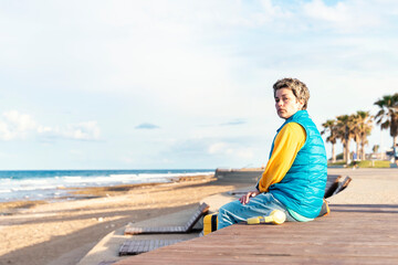 Fototapeta na wymiar Sad Woman in a blue jacket with a yellow suitcase and headphone on the seaside on hot sunny day arrived in a tourist town. Travel Lifestyle concept