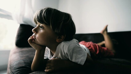 Obraz premium One small boy lying on couch with hand in chin hypnotized by entertainment media at home indoors. Lifestyle shot of kid watching cartoon