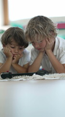 Two small boys watching media on screen laid on floor with hands on chin. Children consuming content online close up faces in Vertical Video