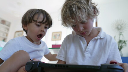 Two small boys looking at video game screen tablet. Younger brother watching older sibling play games on joystick console. Kids playing with technological device