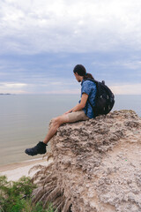 Young latin long haired man with a backpack sitting on a rock while looking at the beach. Vertical...