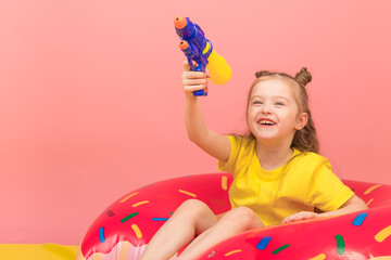 Obraz na płótnie Canvas Cheerful little girl wearing yellow t-shirt and shorts on pink background, lying on a bright inflatable circle and playing with a water gun