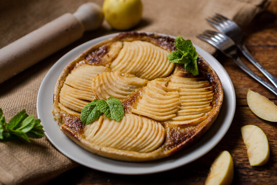 a mouth-watering image of apple French pie placed on wooden surface. Cake is decorated with fresh mint leaves, adding fragrant freshness to it. On crispy dough lined with thin slices of juicy apples, 