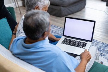 Biracial senior couple using laptop while relaxing on sofa in living room, copy space