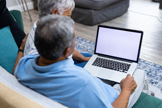 Biracial senior couple using laptop while relaxing on sofa in living room, copy space - Powered by Adobe