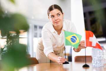 Positive young woman putting little flag of Brazil on the table with flag of Switzerland in...