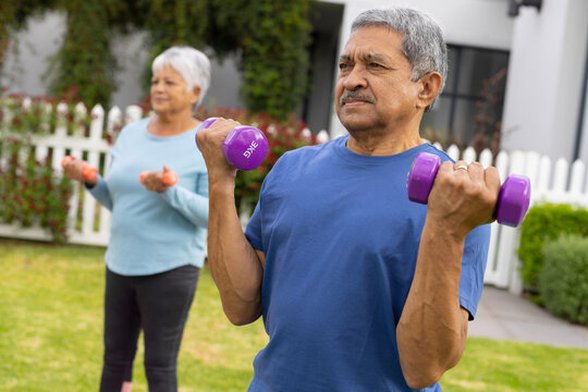Biracial senior couple lifting dumbbells while standing and exercising in yard
