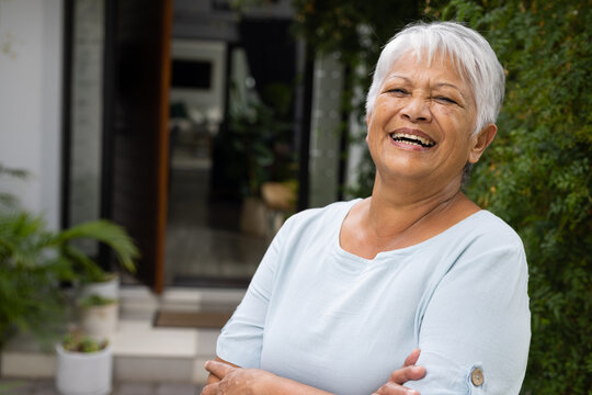 Cheerful biracial senior woman with short gray hair crossing arms and standing outside house