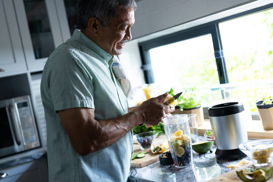 Side view of smiling biracial senior man cutting slices of banana in blender on kitchen counter