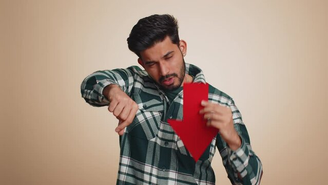 Upset indian man showing red arrow pointing down, concept of downgrade, unsuccessful business, fall of stock market money exchange rate, bankruptcy fail. Hindu guy isolated on beige studio background