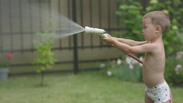 little child boy kid watering plants in garden outdoors in sunny hot summer day. baby child holding watering hose spraying water into green plants, helping gardening. developing assistance, labor 
