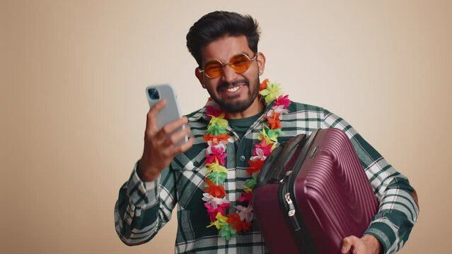 Young tourist indian man with hawaiian lei in sunglasses using smartphone celebrating winning holiday resort vacation tickets pass. Tourism, summer trip to sea. Handsome hindu guy on beige background