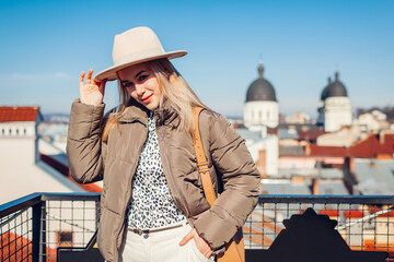 Fashionable young woman enjoying architecture from balcony of European city wearing hat. Spring fashion