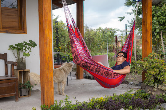 Latino man reclining in a red hammock working on his laptop with a dog accompanying him