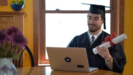 Young man dressing in graduation costume talking to someone online on a laptop showing his diploma.