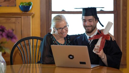 Mother and son sitting hugging at dining room table doing online graduation ceremony concept in costume and holding diploma out to camera.