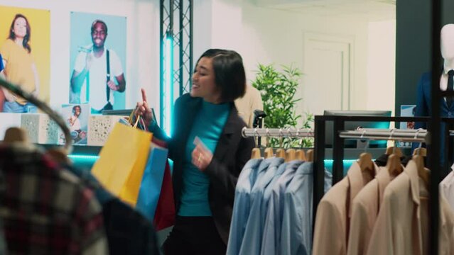 Asian client doing dance moves and feeling silly, leaving with trendy casual wear from fashion boutique cash register. Happy smiling woman dancing with shopping bags, shopping center.