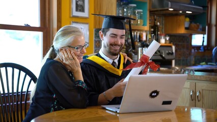 In front of a laptop computer in a home dining room, a mother and her son in graduation attire talk and wave to someone they are teleconferencing with.