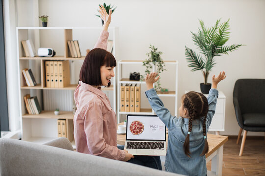 Happy young mother and her cute daughter ordering pizza in internet using modern laptop and cheerful celebration with hands up. Caucasian woman and little girl enjoying time at home with no cooking.