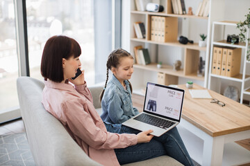 Adorable little girl sitting on couch with her young mother while buying new clothes with discount in internet. Modern caucasian family using smartphone and laptop for online purchase.