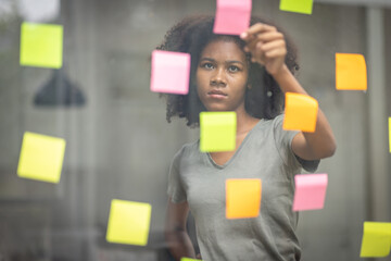 A black American african business woman in afro hairstyle planning in meeting at office and use post it notes to share idea. Brainstorming concept. Sticky note on glass wall.