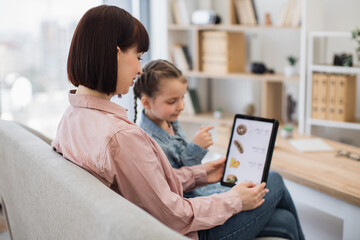 Close up of smiling shoppers choosing fast food at discount on delivery webpage while relaxing at home. Serene woman and girl spending quality time together while buying precooked meals via tablet.