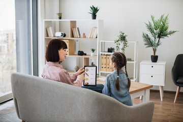 Back view of attractive woman and cute kid surfing takeaway food application on digital device while sitting in elegant lounge at home. Beautiful young ladies ordering restaurant meals using tablet.