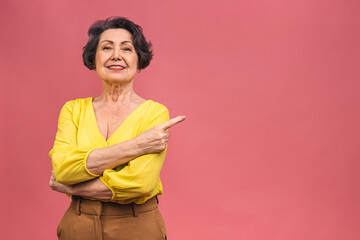 Portrait of attractive cheerful beautiful senior mature aged woman demonstrating copy space isolated over pink background, pointing finger away.