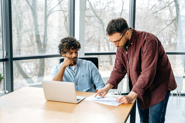 Two male business partners of different nationalities working in a creative modern office, brainstorming on a new project, discussing ideas and strategy, concentrated analysing financial graphs