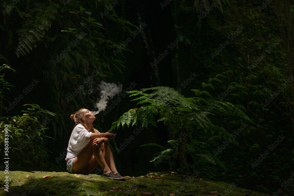 Wall mural young woman smokes a vape in the jungle on a tropical island, bali.