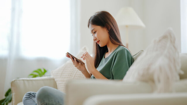 Happy young asian woman relax on comfortable couch at home texting messaging on smartphone, smiling girl use cellphone chatting, browse wireless internet on gadget, shopping online from home