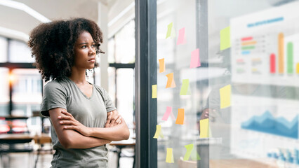 Serously ethnic woman standing with arms crossed looking at sticky note on glass wall, thinking and planning business strategy.