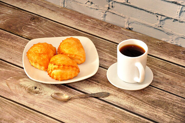 A cup of hot black coffee on a wooden table, next to a plate with three cottage cheese buns.