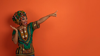 Emotional black lady in african costume pointing at copy space