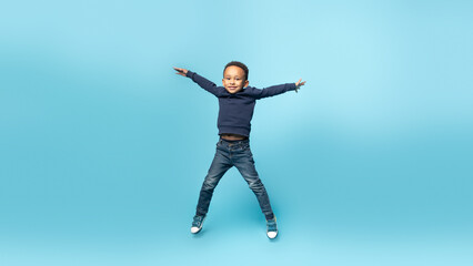 Energetic black boy jumping and raising hands up, having fun and fooling around on blue background, full length