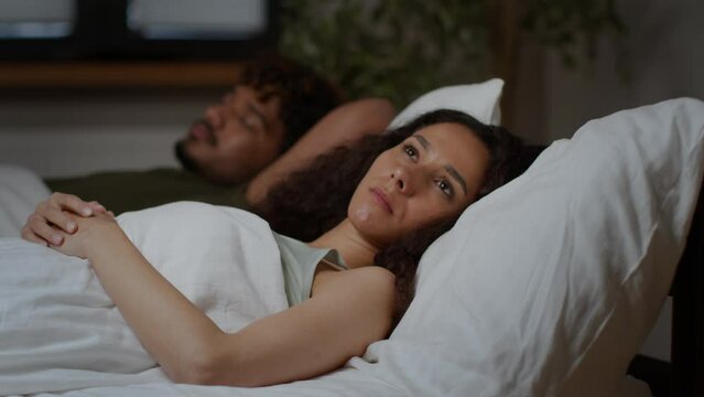 Insomnia. Young middle eastern woman suffering from sleep disorder, lying near sleeping husband in bed with open eyes