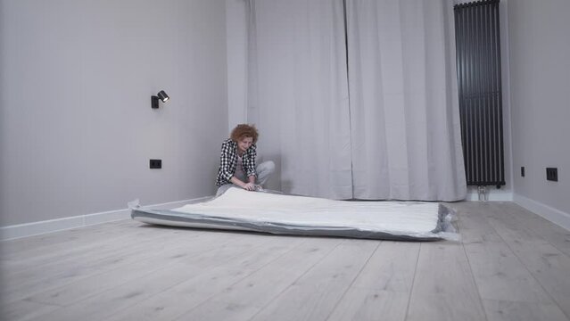 Mature woman unpacking new bed mattress while moving into new apartment. Happy elderly woman unpacking new orthopedic mattress in an empty living room. Furnishing new house. Renting a house. 