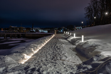 City embankment with walkway covered with snow illuminated of streetlights. Night cityscape.