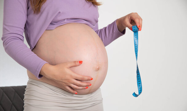 A pregnant girl with a big belly holds a measuring tape on a white background. The concept of measurement and size of the abdomen during pregnancy. The volume of the abdomen by week.