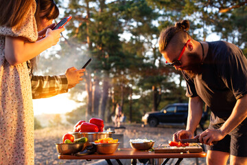 A young man with a beard prepares a salad of vegetables in nature, the girl takes pictures on the phone. Rest friends in the summer in nature, travel, tourism as a hobby.