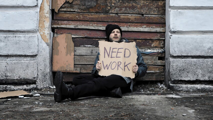 Homeless man sits and holds piece of cardboard with inscription Need Work. Immigrant. Below poverty...