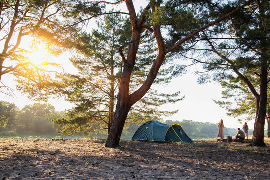 Tourist tent in a picturesque place on the banks of the river against the backdrop of a sunset in summer. A group of friends are resting in the forest in nature. Copy space for text