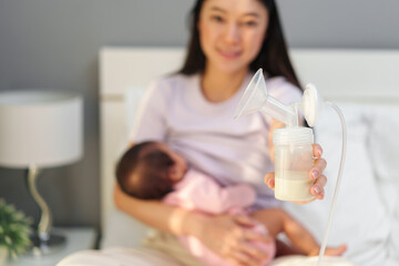 mother holding breast pump milk in bottle while her breastfeeding newborm baby