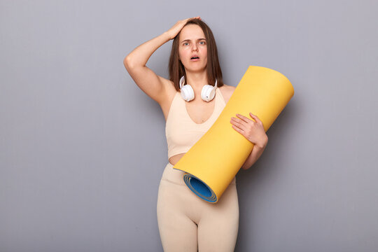 Portrait of shocked scared surprised woman wearing sportswear holding yoga mat, has headphones over neck keeps hand on her head, looking at camera with big eyes, standing isolated over gray background