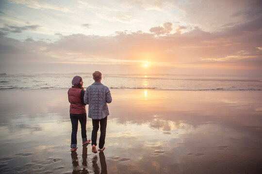 Rear view of couple walking at beach against sky during sunset