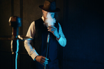 Front view of resting man smoking hookah at home workplace with neon lighting