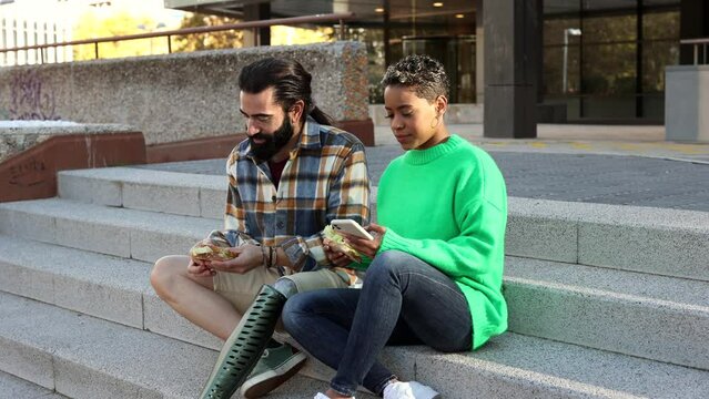 Traveling multiracial couple are eating in the city outdoors sitting on some stairs, man with amputated leg wears a prosthesis.