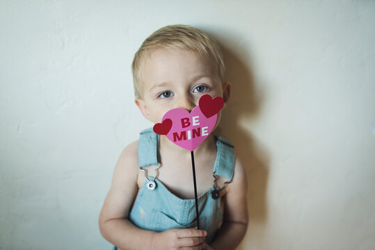 Portrait Of Cute Boy Holding Heart Shape With Be Mine Text Against Wall