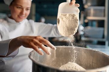 Female confectioner wearing white uniform putting flour into big metal bowl