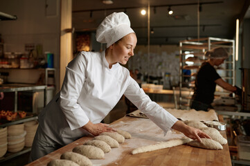 Woman baker forming bread loaves from raw dough at professional kitchen - obrazy, fototapety, plakaty